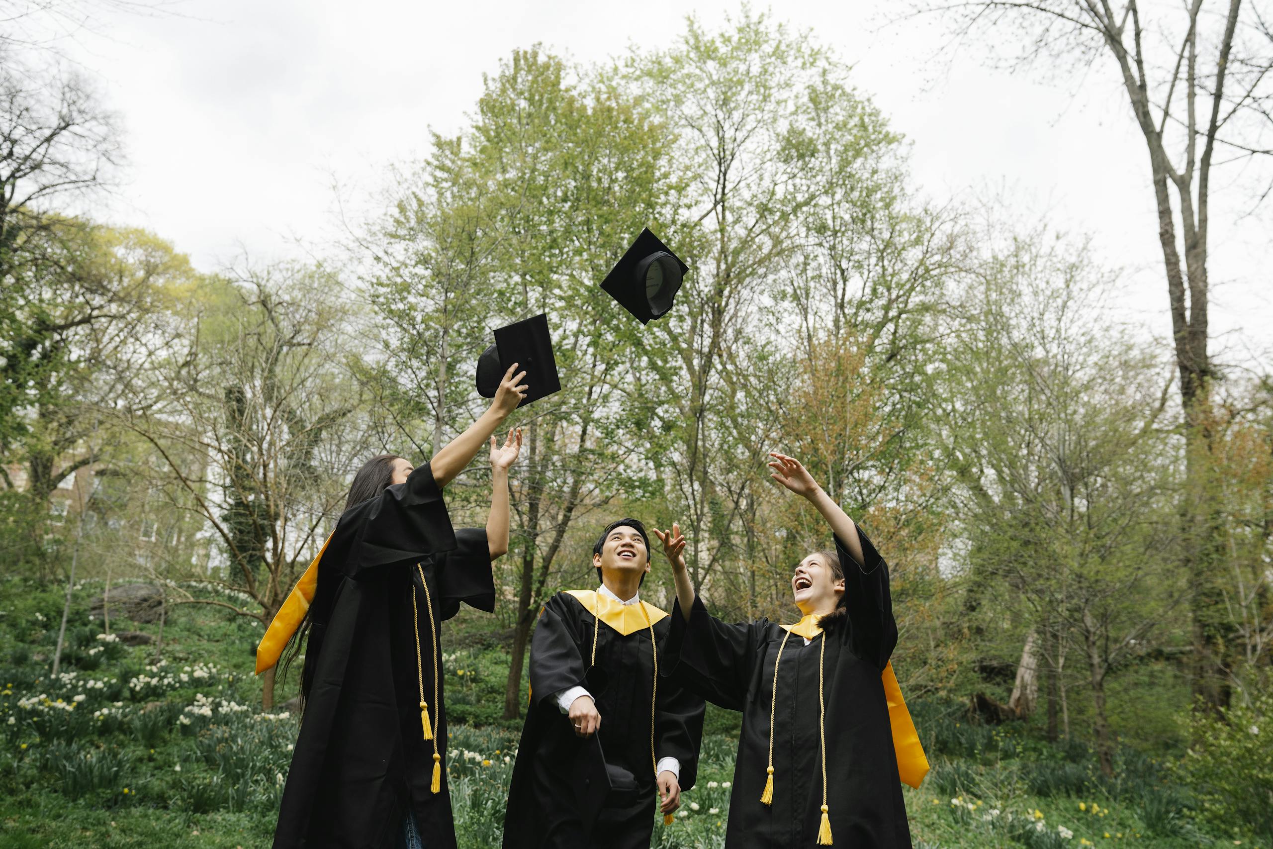 Three graduates celebrating and throwing caps in the air in a lush green park.